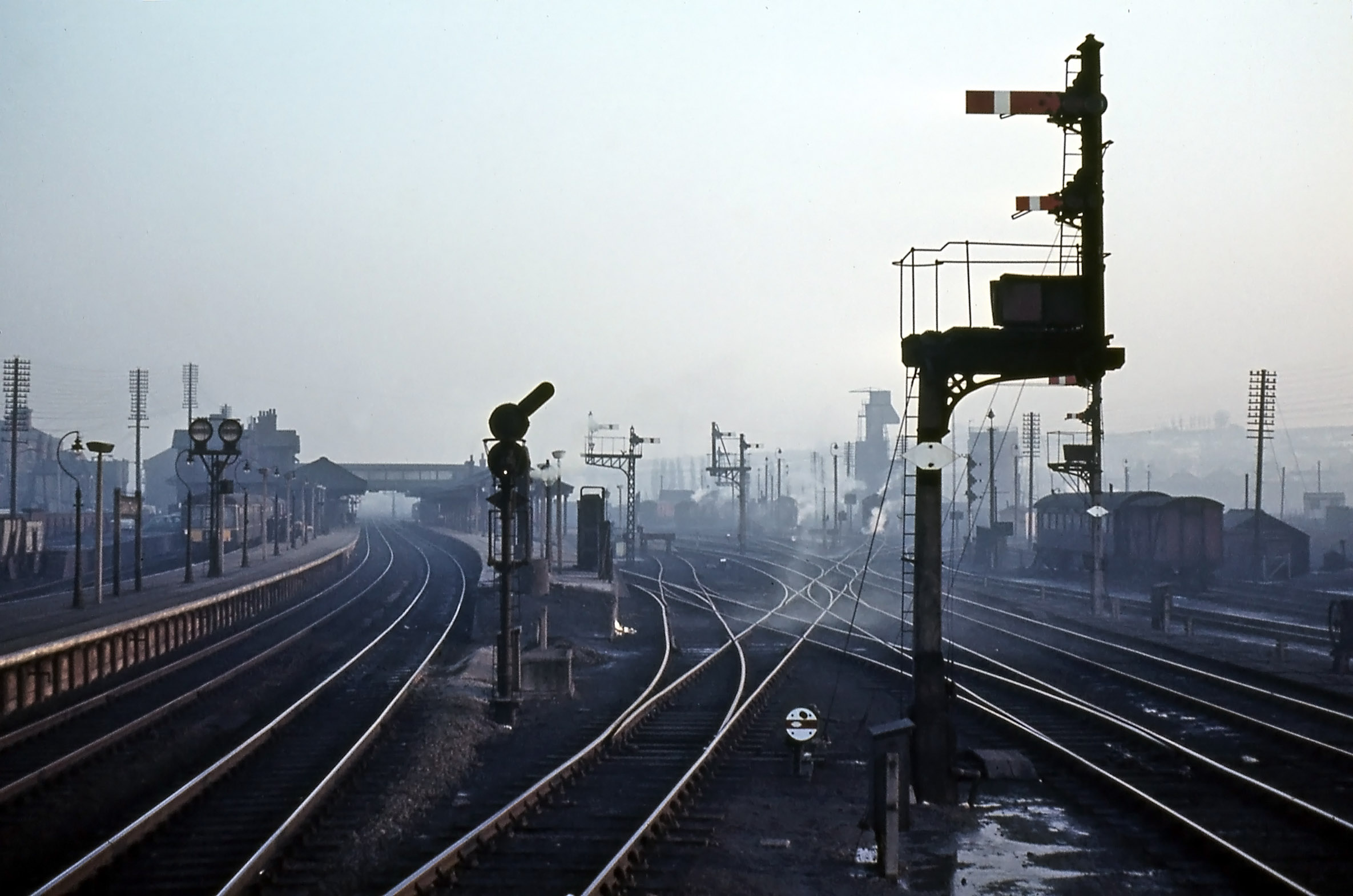 This photograph was taken in 1962 from about halfway up the signalbox steps. Compared with the previous view: o The tall Main line signals have been replaced with a colour light signal which showed an inclined row of white lights if the Nottingham line was to be taken. o The Bay platform and Western platform departure signals are unchanged. o The old-style signal controlling access from the Nottingham direction has been replaced by the bracketed post in the right foreground. It has one full-sized and one miniature arm together with an electric route indicator in the box beneath them. The full-size arm gave access to one of three principal routes, provided that it was clear. The miniature arm permitted access to sidings or, under caution, to a principal route which was already occupied. When either arm was raised the route indicator showed a code to inform the driver of the route set: o DB – Down Bay o ES – Engine Spur (miniature arm only) o W – Western platform o C – Carriage sidings (miniature arm only) o UG – Up Goods line o S – Shunt line (miniature arm only) Further back and across to the right is a similar signal post controlling access to the Goods line in the southbound (or Up) direction. Again it has both a full size and a miniature arm, and a route indicator capable of showing routes as follows: o C – Carriage sidings (miniature arm only) o UG – Up Goods line o S – Shunt line (miniature arm only) Beyond is a signal post with three miniature arms mounted vertically. This was the shed outlet signal which is seen more clearly in the next photograph. Photograph by Noel Ingram, used with permission from Steam World.