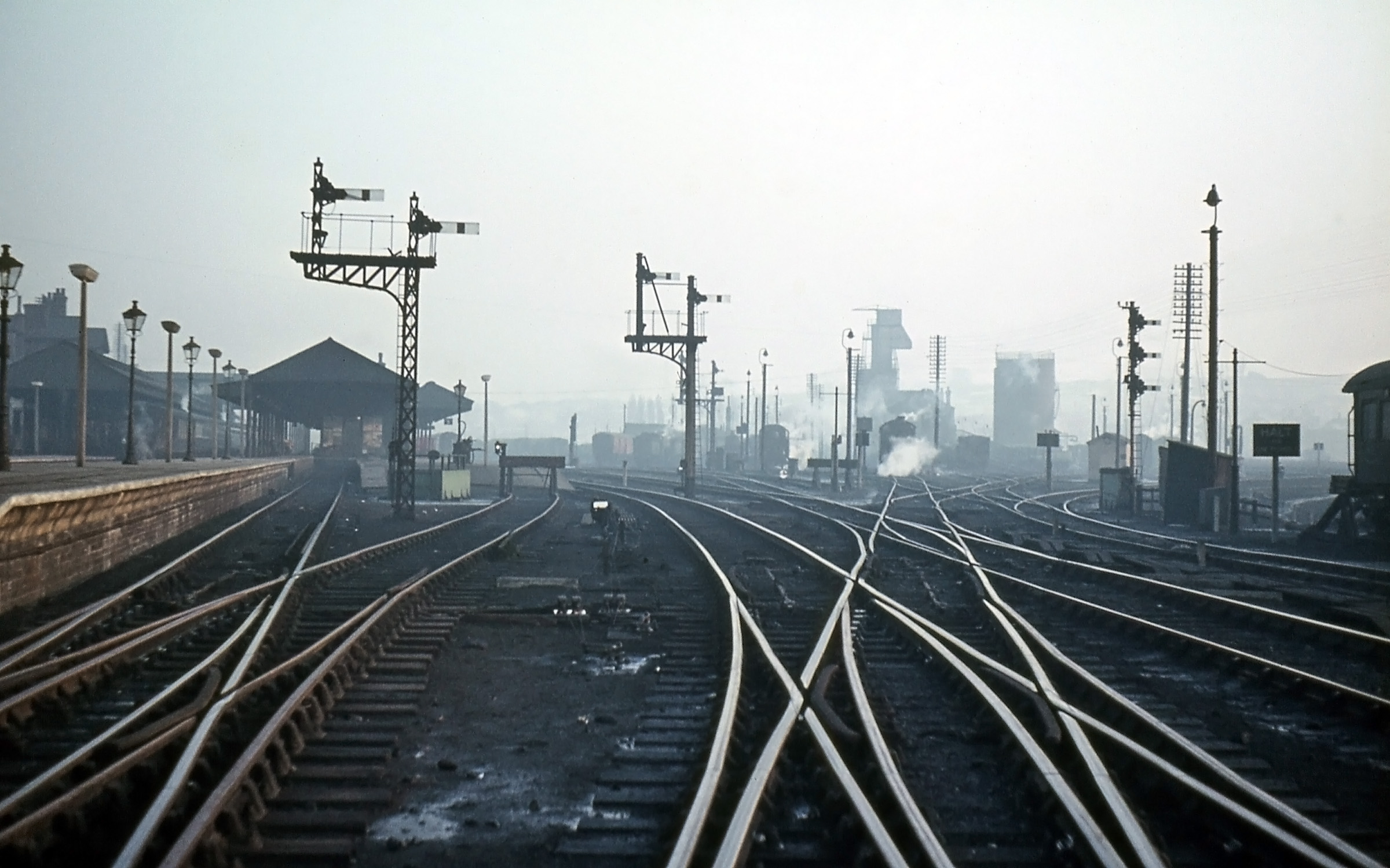 On the same evening in 1962 Noel Ingram has taken a few steps forward to better capture the ‘atmosphere’ of the shed and the glint of the clear evening sky on polished steel rails. The shed outlet signal is prominent on the right. Reading as before ‘top to bottom’ = ‘left to right’ the top arm indicated the Goods line, the middle arm the Nottingham line and the bottom arm the Main line. Nearby there was a box containing a telephone from which the fireman of every locomotive leaving the shed would phone the North Box signalman to tell him which train they were for, so that the correct road could be set. If they were going to stand ready to take forward an incoming train the signalman would relay information about how it was running. The elegant cast iron gas lighting columns on the platform are being replaced by electric lights on concrete posts. In the distance at the centre, partly obscured by the Western platform departure signal, is another bracket signal which controls the Up & Down Goods line in the Down direction – see the next photograph. Photograph by Noel Ingram, used with permission from Steam World.