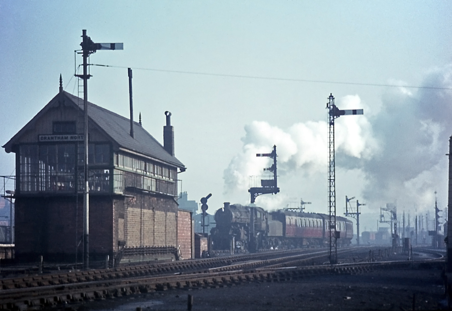 On Monday 25th February 1963 a mid-afternoon service for Lincoln, hauled by 4MT locomotive No. 43146, is departing from the Western platform, using the connection between the end of the down side platforms and Grantham North signal box to join the Down Main line. The four signals nearest the photographer in this view are, from left to right: o the signal for trains departing on the Nottingham line (nearest the signal box) o the platform starting signal for the Down Main line (a colour light signal) o the signal for trains arriving from the Nottingham line (nearest the signal box) 0 the signal for trains departing for the north on the Goods line (to the right of the Nottingham line) As evidenced by the stove pipe and the chimney, due to its length Grantham North box had the 'luxury' of two coal-fired stoves. Photograph by Noel Ingram, used with permission from Steam World.