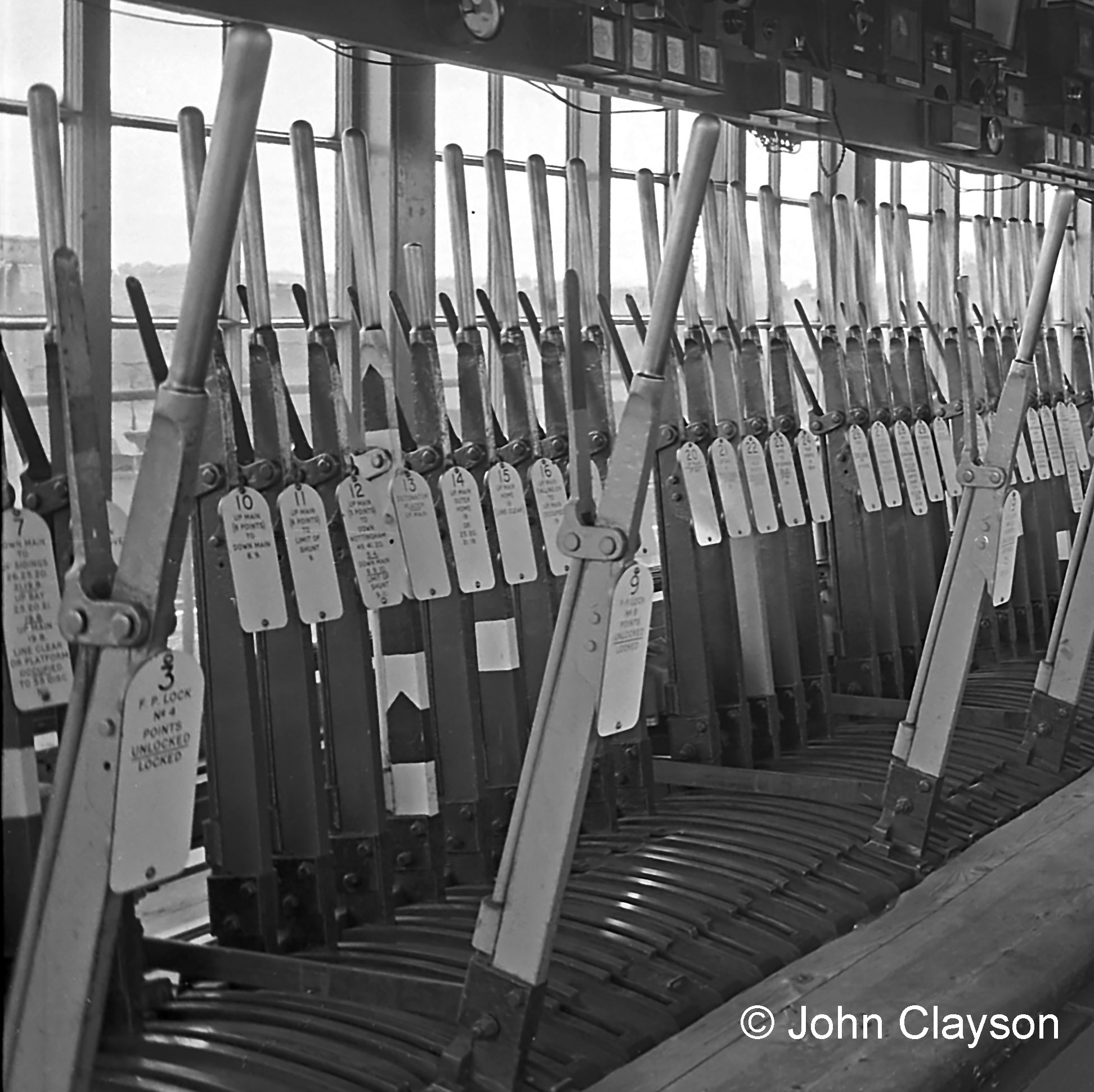 Here is the north end of the lever frame at Grantham North box on 22nd August 1963. Levers in a signal box are all numbered, and also painted in colours according to their purpose. For those of us who are viewing in black and white (as snooker commentator Ted Lowe might say), the red levers (e.g. 10, 11, 12, 14, 15 & 16) control stop, calling-on or shunt signals, the black levers (e.g. 20 & 21) are for points, and the blue levers (e.g. 3, 9, 19 & 22) are for facing point locks (which are a safety device to lock points in place once they are set). The lever with black and white stripes and a chevron, 13, is for a detonator placer on the Up Main line – an emergency device that places a small explosive charge (detonator) on the rail. The detonator explodes when run over by a train’s wheels, warning the driver of danger. The information on the lever plates tells us that levers in this part of the frame control some of the signals and points relating to the Up Main line seen in the photograph next above. For example: 15 UP MAIN HOME is the topmost of the signals which the train is passing; the white band on the lever shows that it is interlocked electrically with the block telegraph (as well as mechanically in the lever frame). 16 UP MAIN CALLING-ON TO UP MAIN OCCUPIED is the small signal on the same post, beneath the distant signal. Moving closer to the camera, the two disc signals visible against the third coach are (left to right): 10 UP MAIN TO DOWN MAIN (relates to a trailing crossover hidden under the train). 11 UP MAIN TO LIMIT OF SHUNT. Closer still: 19, 20, 21 & 22 control the points for the connection to the Up bay platform, in the right foreground. Photograph by Cedric A. Clayson.