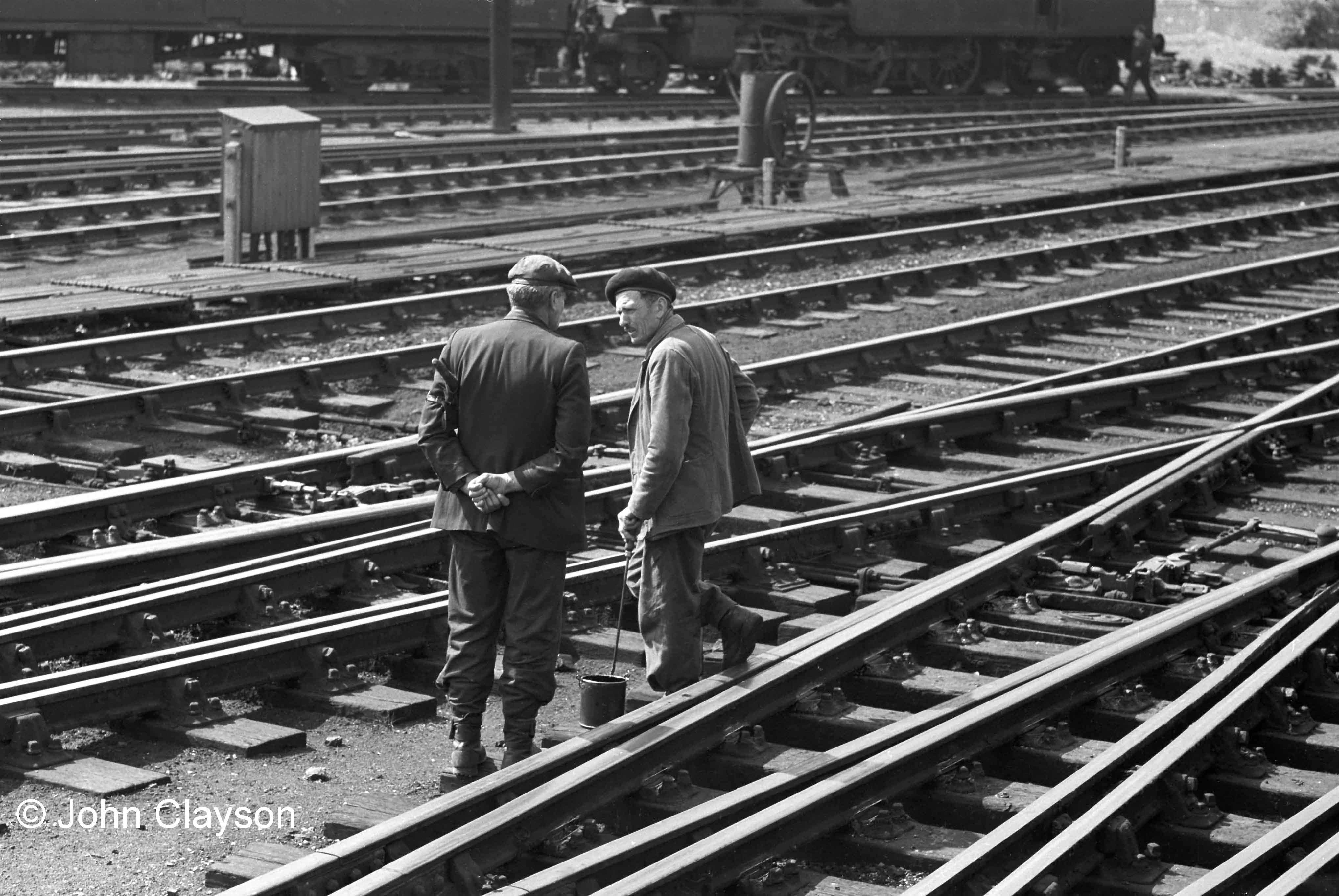 The man on the right is Augustus (Gus) Summers, a former German PoW who worked for Tearson’s (Terson’s?), a contractor who carried out track maintenance and repairs. With his pot of oil and long-sticked brush he is lubricating the point mechanisms near the north end of the station. If you look at the track in the foreground you can see that there is fresh oil around the special rail chairs fixed to the crossing timbers, known as 'slide chairs', on which the switch blades slide. The man on the left is Tom Plummer, the North End Ganger at Grantham, who is acting as ‘Look-Out’ – he is carrying a set of flags under his left arm, on which he also has strapped an enamelled ‘Look-Out’ armband. This was, and still is, a vital role - keeping watch for, and giving warning of, approaching trains which might not be noticed by track workers engaged in a maintenance task on lines which remain open to traffic. The regular lubrication of points is important because the lengths of rail which move across the slide chairs when points are changed are heavy – on a crossover (two points worked together) they might weigh more than a tonne. The signalman’s muscle power also has to move the heavy point rods linking the signal box lever frame with the points. Today, most points on main lines are worked by electric motor. The object in the centre background is a drilling machine, which was used for boring holes in point stretcher bars. May 24th 1962 Photograph by Cedric A. Clayson,