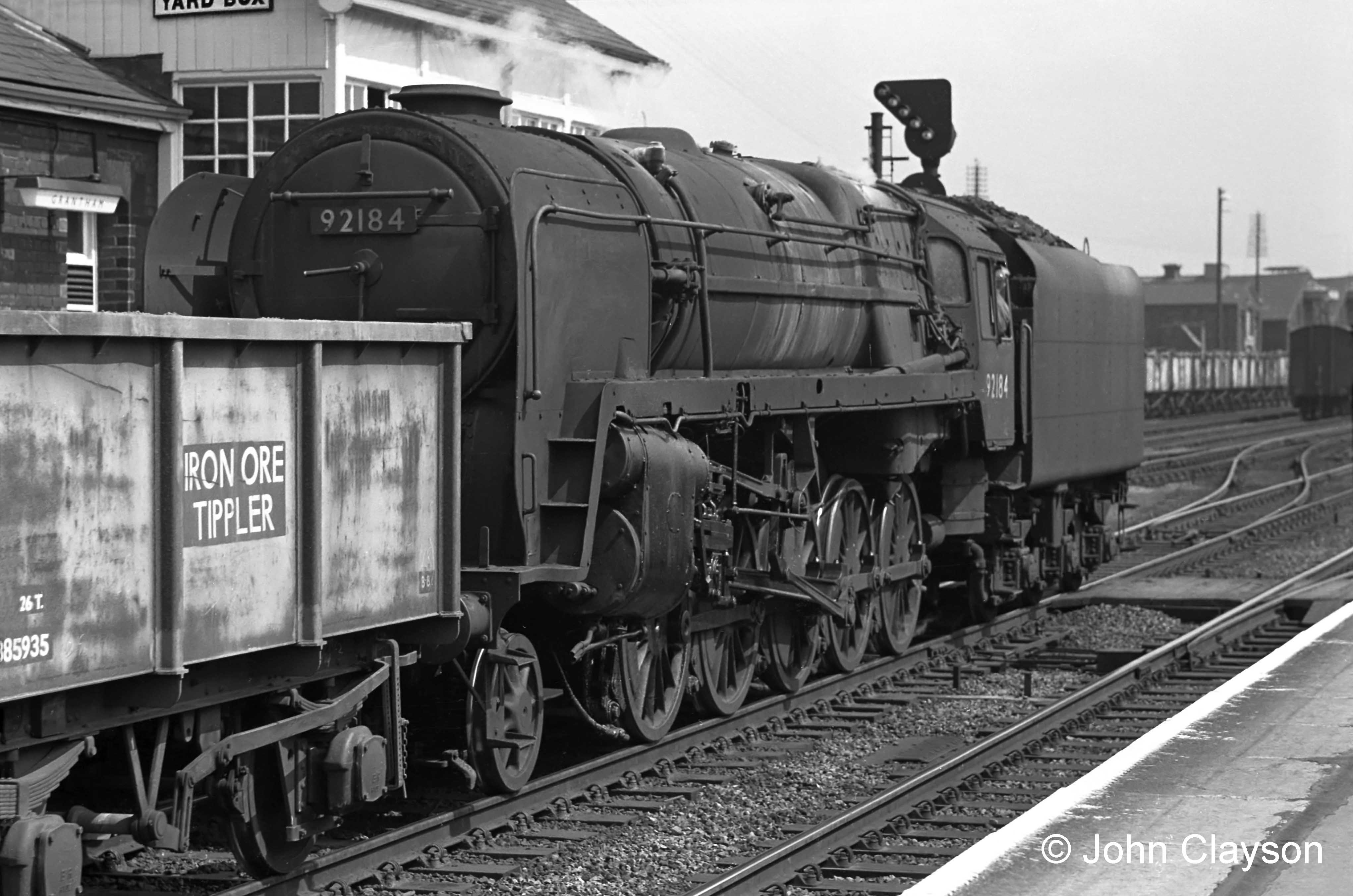 A train of empty ironstone wagons is being taken south through the station on the Up Main line by class 9F locomotive No.92184 on 27th June 1963. Projecting above the level of the cab is an inclined row of illuminated white lights attached to the colour light signal. This shows that the train is signalled to take the connection into the Up Goods line at the points just beyond its tender. It will use the Goods line, alongside the Up Main line, from here as far as High Dyke, where it will be crossed over to the Down side to enter the reception sidings for the Stainby Branch. The Goods line kept slower trains, such as this one, out of the path of the expresses, especially important because the route climbs continually from here to Stoke Tunnel (just beyond High Dyke), slowing mineral trains even more. The term ‘Iron Ore Tippler’ indicates that the wagons have no doors – they were emptied by being tipped upside down in unloading plant at the steelworks. Photograph by Cedric A. Clayson.