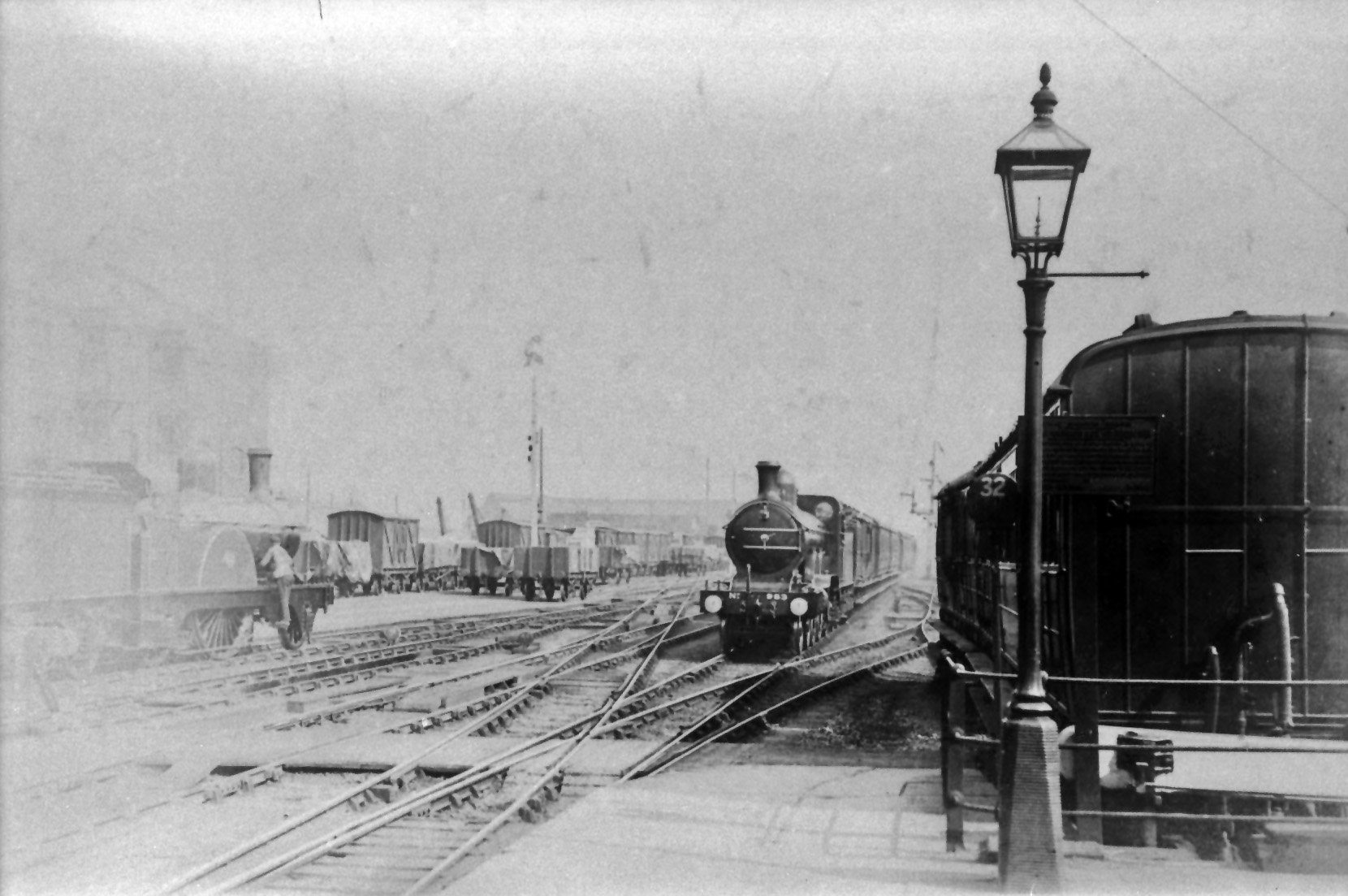 In this view of 17th June 1902 looking south Grantham Yard signal box is just off shot to the left. On the left are the two locomotive spurs, with shallow pits to assist inspection and oiling round, where locomotive No. 877 is waiting to take over a southbound passenger train. To the left of the approaching northbound express is a series of crossings which link, in receding order, the Down and Up Main lines to the Up Goods line and the goods yard sidings. The northbound express is approaching on the Down Main line under clear signals, the upper arm controlled by the Yard Box, the lower by the North Box. In the shadow of the coaches on the right is a spindle-mounted disc signal to control shunting movements from the Down Main platform across the connection to the right. It has '32' picked out in white paint - the number of the lever in the Yard Box which operated it. The dock in which the coaches are standing was one of two short sidings, long-standing features of the layout where coaches or vans could be kept in readiness for adding to northbound trains. Photograph by Rev. T.B. Parley from the John Tatchell Collection of the Historical Model Railway Society (HMRS).