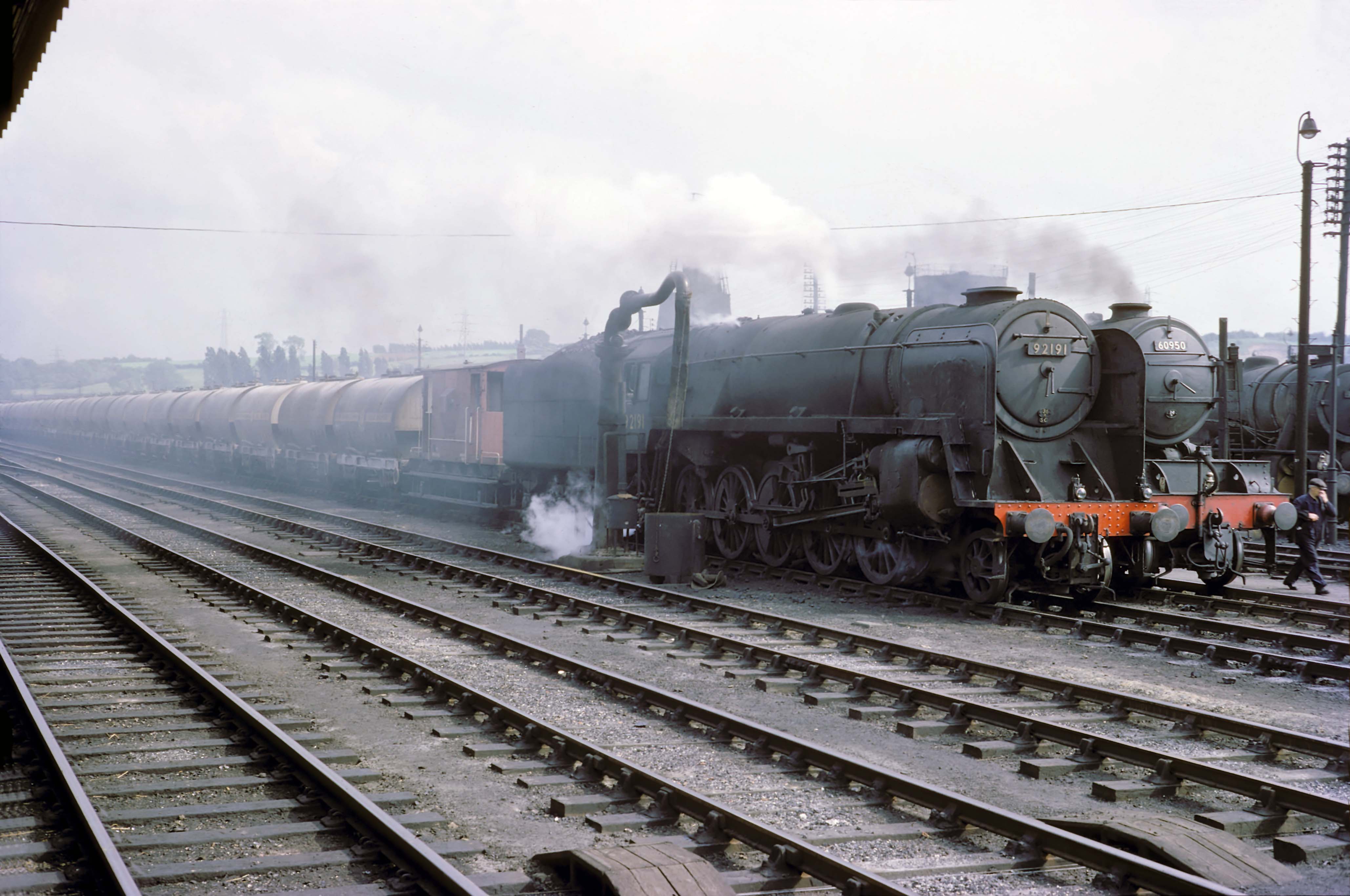Class 9F locomotive No. 92191 of Doncaster (36A) motive power depot is hauling the northbound, loaded, Blue Circle Cement train. It has paused on the Up and Down Goods line west of the station platforms, probably to wait for a ‘path’ between passenger trains. Behind is class V2 No. 60950 of Grantham (34F) shed, also on a northbound freight. Photograph by Cedric A. Clayson, © John Clayson