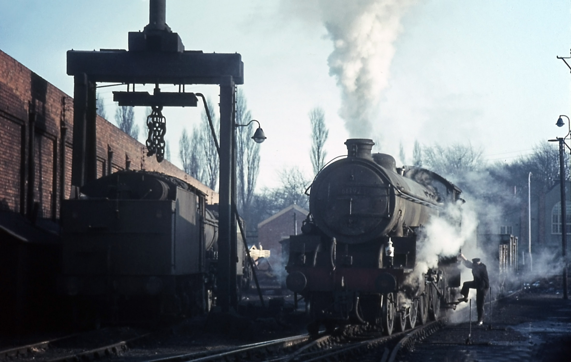 This view is taken in the oposite direction to the one above. Climbing aboard B1 locomotive No.61392 as it accelerates down the yard is driver Jim Willetts whose son, Ken, was also a driver at Grantham. Catching the sun between the two locomotives is the white-painted buffer stop which formed the end of the spur at one of the apexes of the truning triangle. Mick Grummitt, telegraph lad, remembers being at work one day at the North Box when a train came in from the north with a hot axlebox on the locomotive. The locomotive was changed and its crew, from Heaton in Newcastle, went onto the 'angle' to turn. Not being familiar with the layout they failed to stop in time, demolished these buffers and made a hole in the wall of the shed. This rendered the 'angle' out of use until the locomotive was recovered, and engines had to travel to Barkston Junction to turn. Photograph taken by Noel Ingram, used with permission from Steam World.