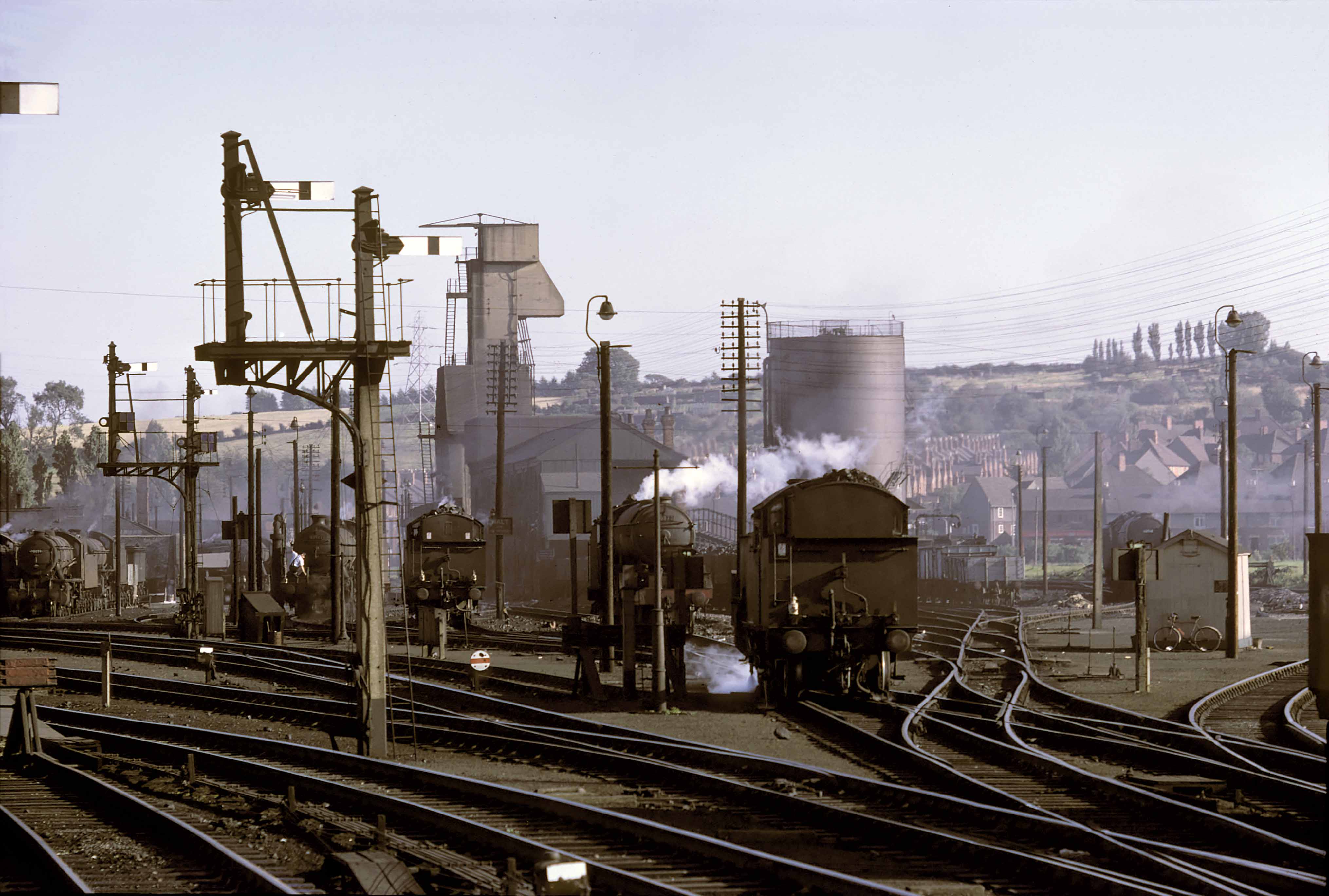 Looking south west from the north end of the down platform on 31st August 1961. Prominent on the skyline is the tall mechanical coaling plant, built of reinforced concrete in 1936-37. Wagons of coal were hoisted in turn to the top and emptied into an internal hopper above the tracks. Locomotives were placed underneath to have their tenders and bunkers replenished. To its right is the tank of the water treatment plant, a little older than the coaling plant, which supplied water for locomotives. The treatment inhibited the formation of scale inside steam locomotive boilers. The cabin near the right of the photograph was the Pilots Cabin, a messroom where the local men who were on main line standby pilot duty would await the call to action, and visiting locomotive crews could rest and make a brew while waiting for their return working. The bike probably belonged to caller-up and odd-job man Pete Ballaam, who would often be sent out to get pork pies from Watkin's Pork Butchers in the town for visiting crews - especially 'Cockneys' as they were referred to (from King's Cross depot) who relished this local speciality. Watkin & Sons closed in 1990 after 78 years in business. The 2 loops near the exit from the loco where prepared engines were placed was called 'London road'. There was a phone there to the North Box to tell the 'bobby' when you were ready to leave the shed. Locomotives of types WD (90059), B1, O2, L1, and V2 (60872 King's Own Yorkshire Light Infantry) can be seen, and there is a fully-coaled A2/3 class pacific loco in the distance, beyond the Pilots Cabin. This is probably the 'standby pilot', or 'main line pilot', an express locomotive which was kept ready in case of a breakdown on the main line. It is standing on one of the lines which was part of the Grantham shed triangle (known to local railwaymen as the angle), so that it could be turned quickly (taken round the 'angle) if required to assist with a southbound train. Photograph by Cedric A. Clayson, © John Clayson 